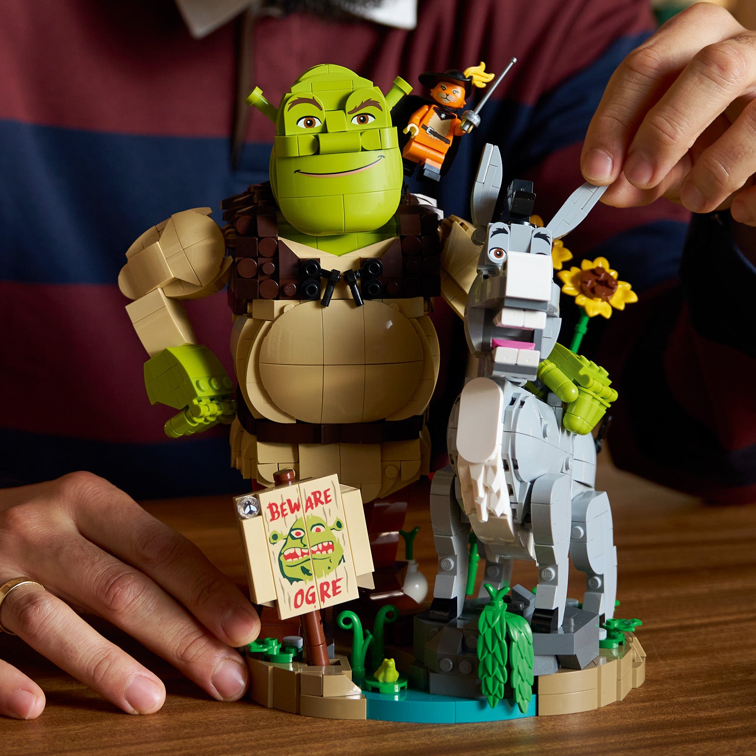 LEGO Figuren von Shrek, Esel und dem gestiefelten Kater auf einer kleinen Landschaft mit Wasser und Pflanzen, gehalten von einer Person im Hintergrund