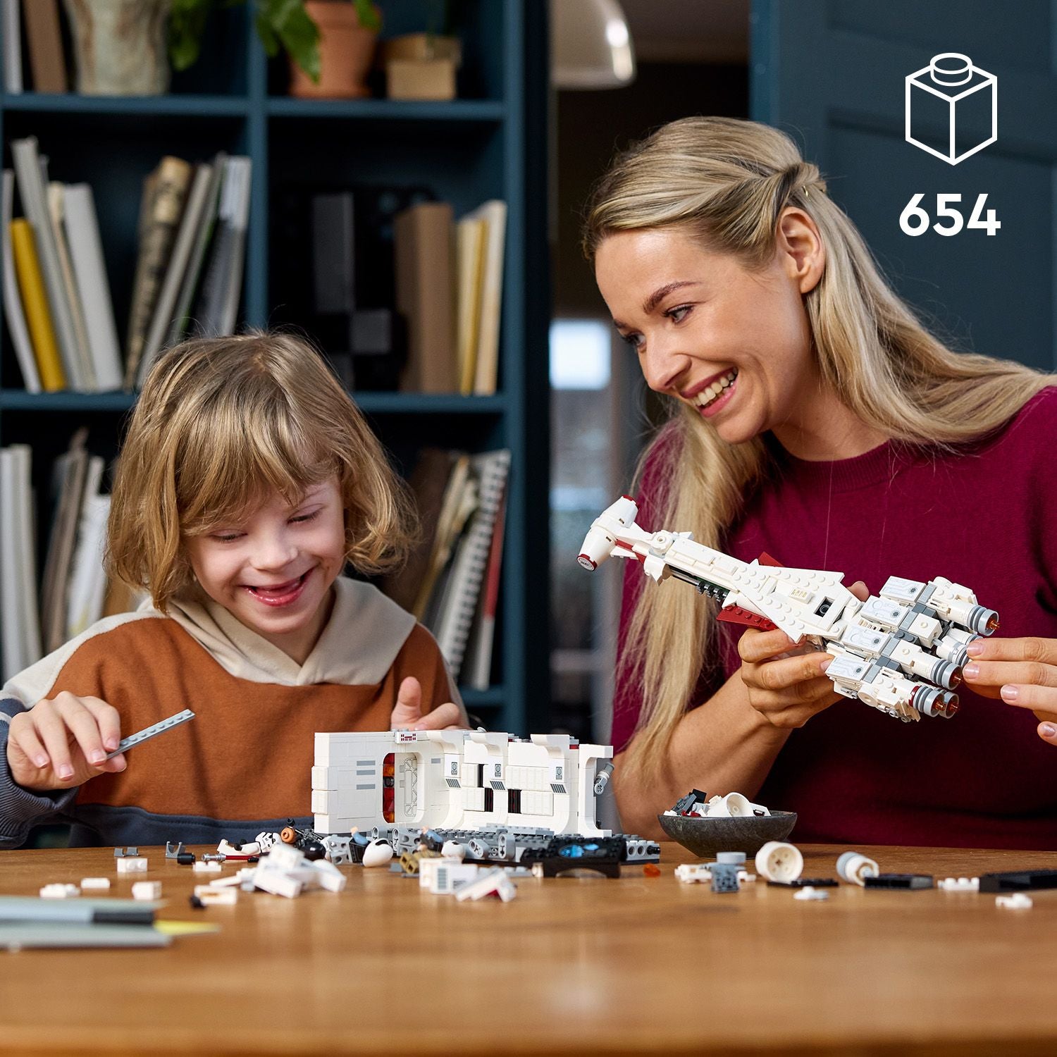 Zwei Personen bauen am Tisch ein weißes LEGO Star Wars Raumschiffmodell mit vielen Einzelteilen, darunter ein länglicher Rumpf und runde Triebwerke.