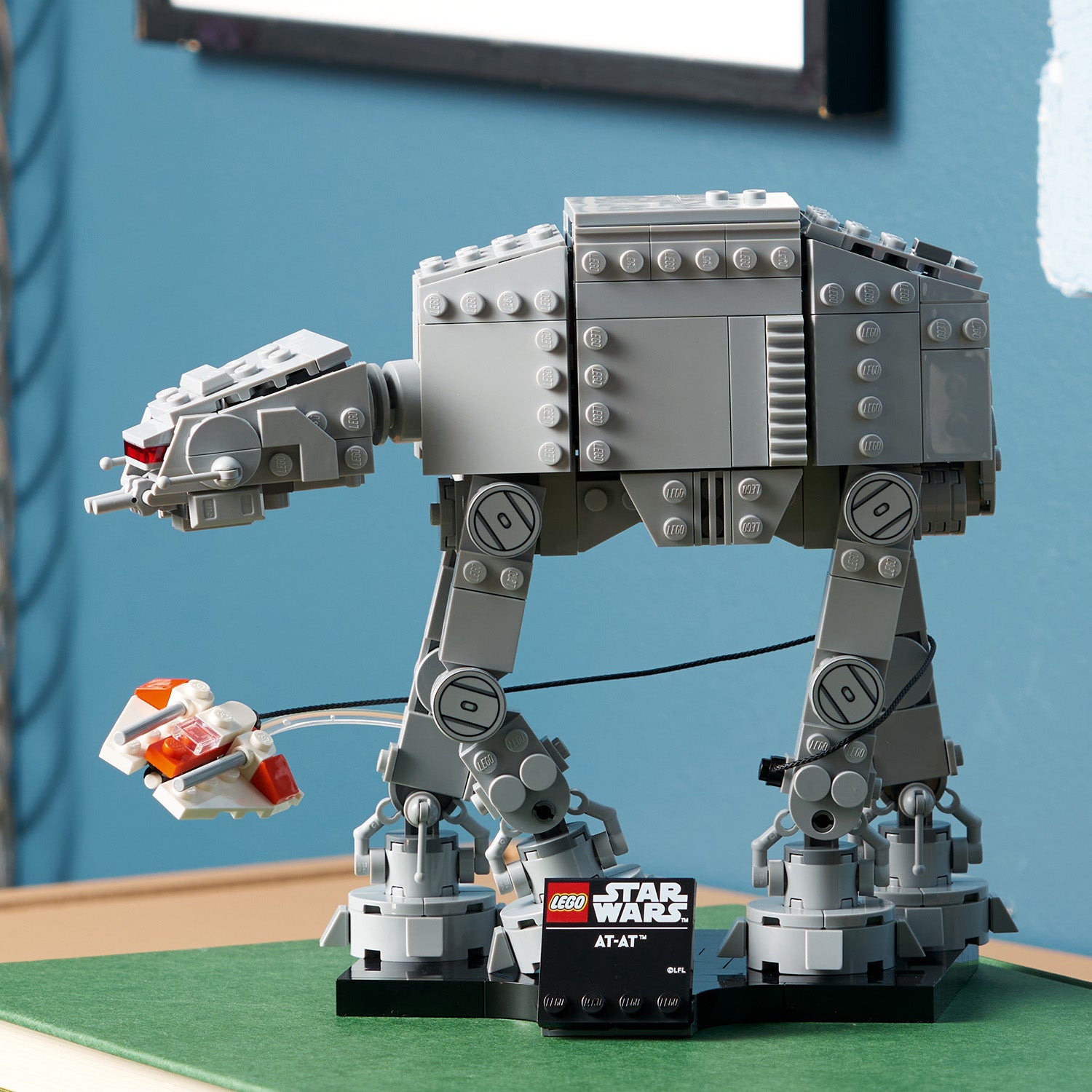 Graues LEGO Modell eines AT-AT aus Star Wars mit beweglichen Beinen auf grünem Untergrund, daneben ein kleines LEGO Raumschiff und ein schwarzer Sockel mit dem Schriftzug 'LEGO Star Wars AT-AT'.
