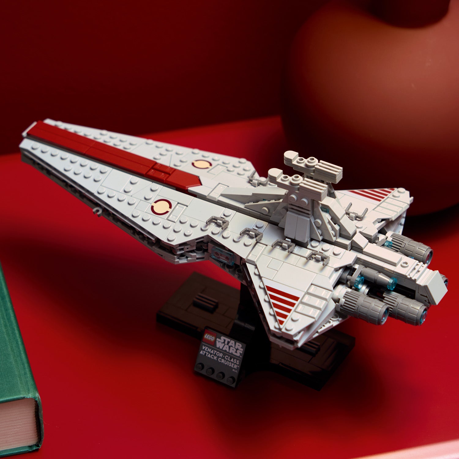 LEGO Modell des Venator-Klasse-Angriffskreuzers aus Star Wars, auf einem schwarzen Ständer mit Namensschild, auf rotem Untergrund neben einem grünen Buch.