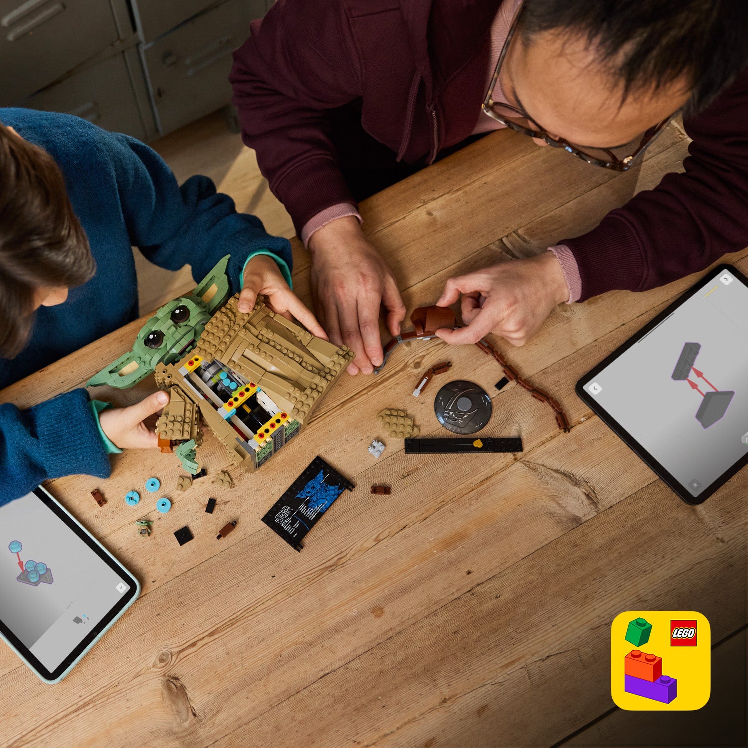 Zwei Personen bauen an einem LEGO Star Wars Set mit Grogu-Figur auf einem Holztisch, daneben liegen LEGO Steine und zwei Tablets mit Bauanleitungen.