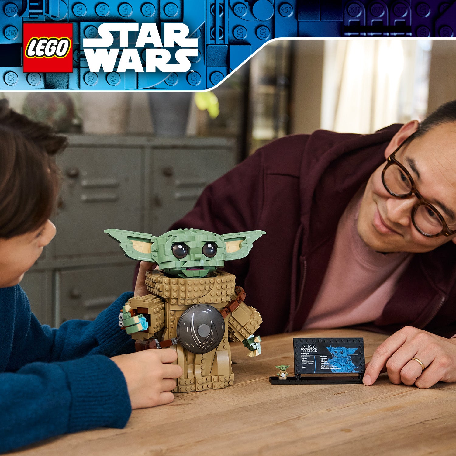Zwei Personen spielen mit einer LEGO Star Wars Grogu-Figur auf einem Holztisch, daneben steht ein kleines Display mit einer Bauanleitung oder einem Datenblatt.