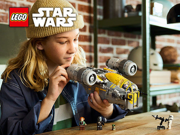 Kind hält das LEGO Star Wars Modell Razor Crest in den Händen, im Hintergrund ein Regal mit Büchern und Dekoration.