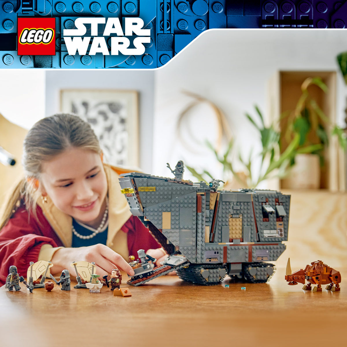 Mädchen spielt mit LEGO Star Wars Sandkrawler™-Modell und mehreren Minifiguren auf Holztisch in hellem Raum