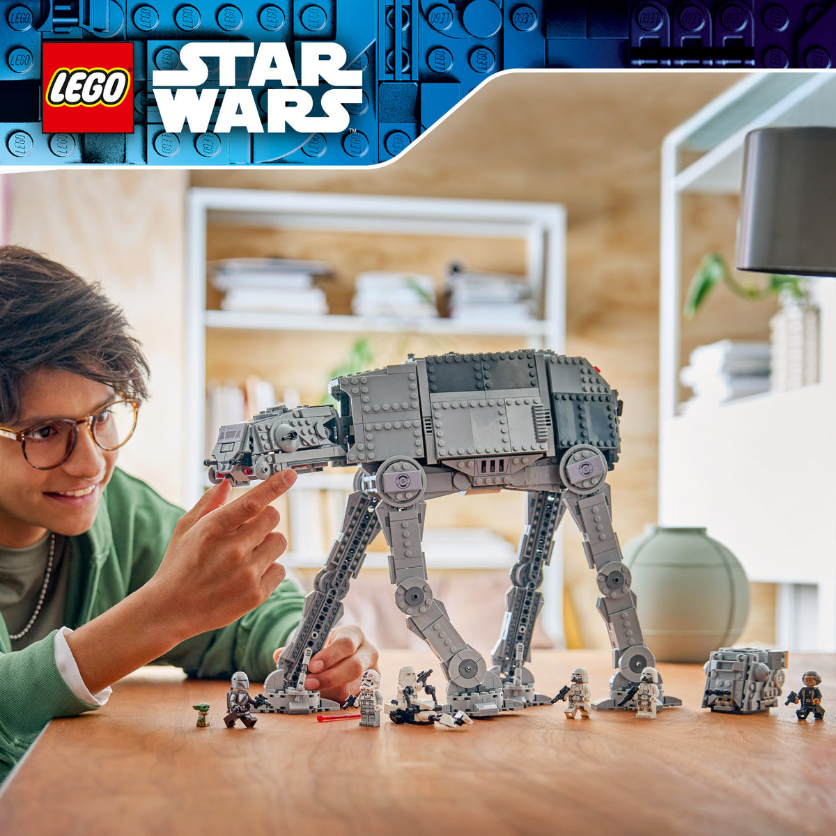 Junge spielt mit einem grauen LEGO Star Wars AT-AT-Modell auf einem Holztisch, umgeben von mehreren kleinen LEGO Figuren und Objekten in einem hellen Raum mit Regalen im Hintergrund