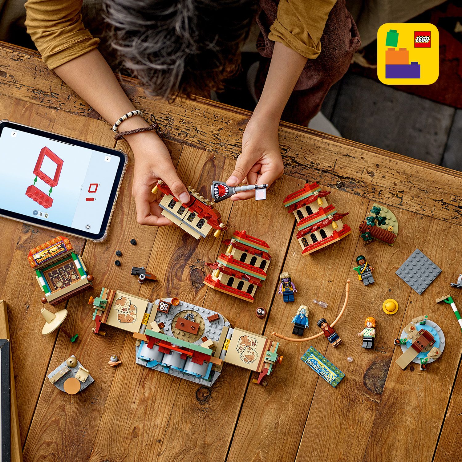 Person baut mit LEGO Steinen ein mehrteiliges Gebäude auf einem Holztisch, daneben Tablet mit Bauanleitung und mehrere LEGO Figuren.