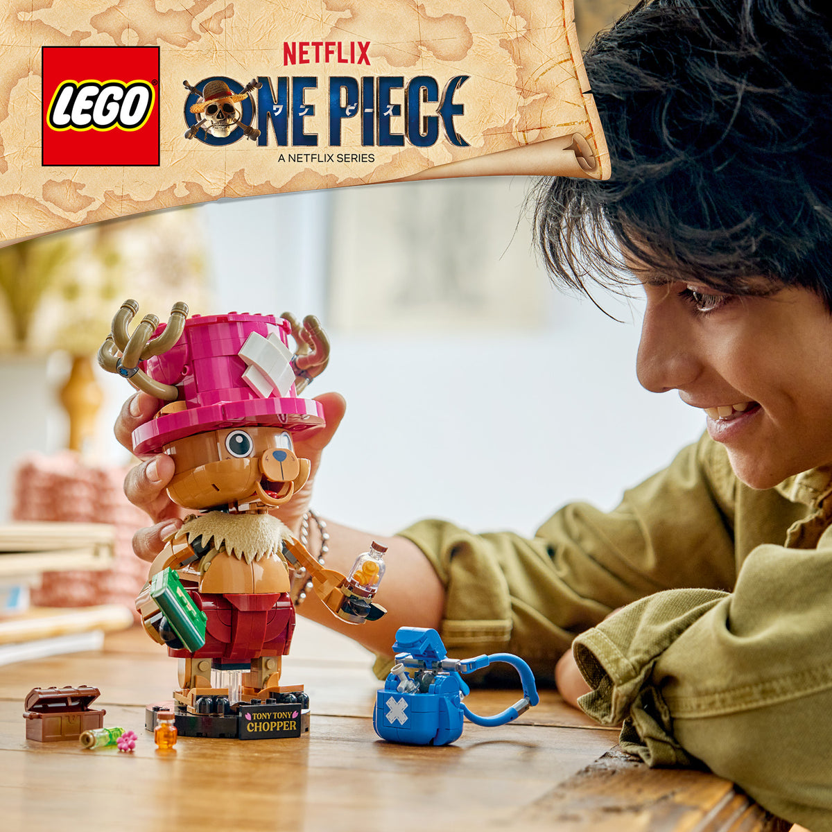 LEGO Figur Tony Tony Chopper aus der Serie One Piece mit rosa Hut und roten Shorts steht auf einem Tisch neben einem blauen Beutel, ein Junge schaut lächelnd darauf.