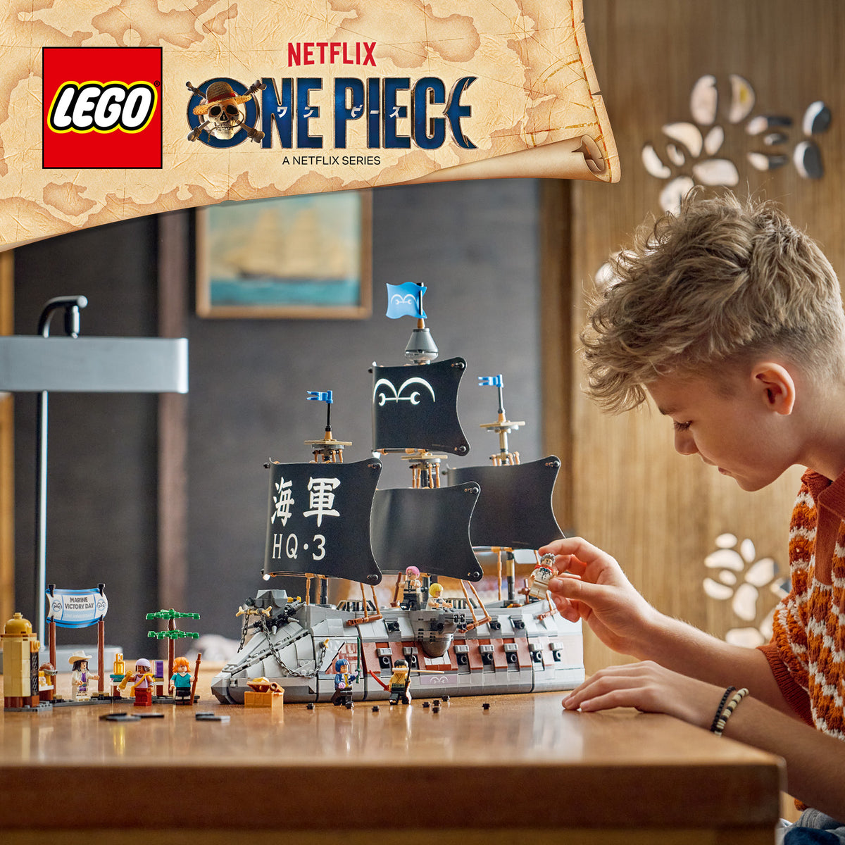 Ein Junge baut an einem LEGO Schiffsmodell mit schwarzen Segeln und asiatischen Schriftzeichen, umgeben von LEGO Figuren und Zubehör, auf einem Holztisch in einem hellen Raum.