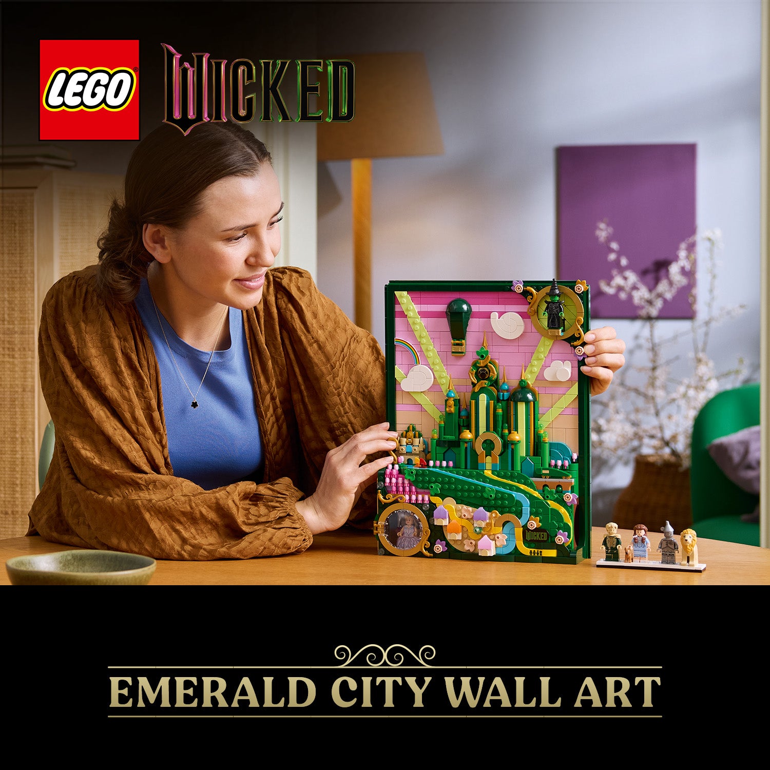 Person hält LEGO Wandkunst mit Emerald City-Motiv aus dem Wicked-Set, umgeben von Minifiguren auf einem Tisch in einem Wohnzimmer.