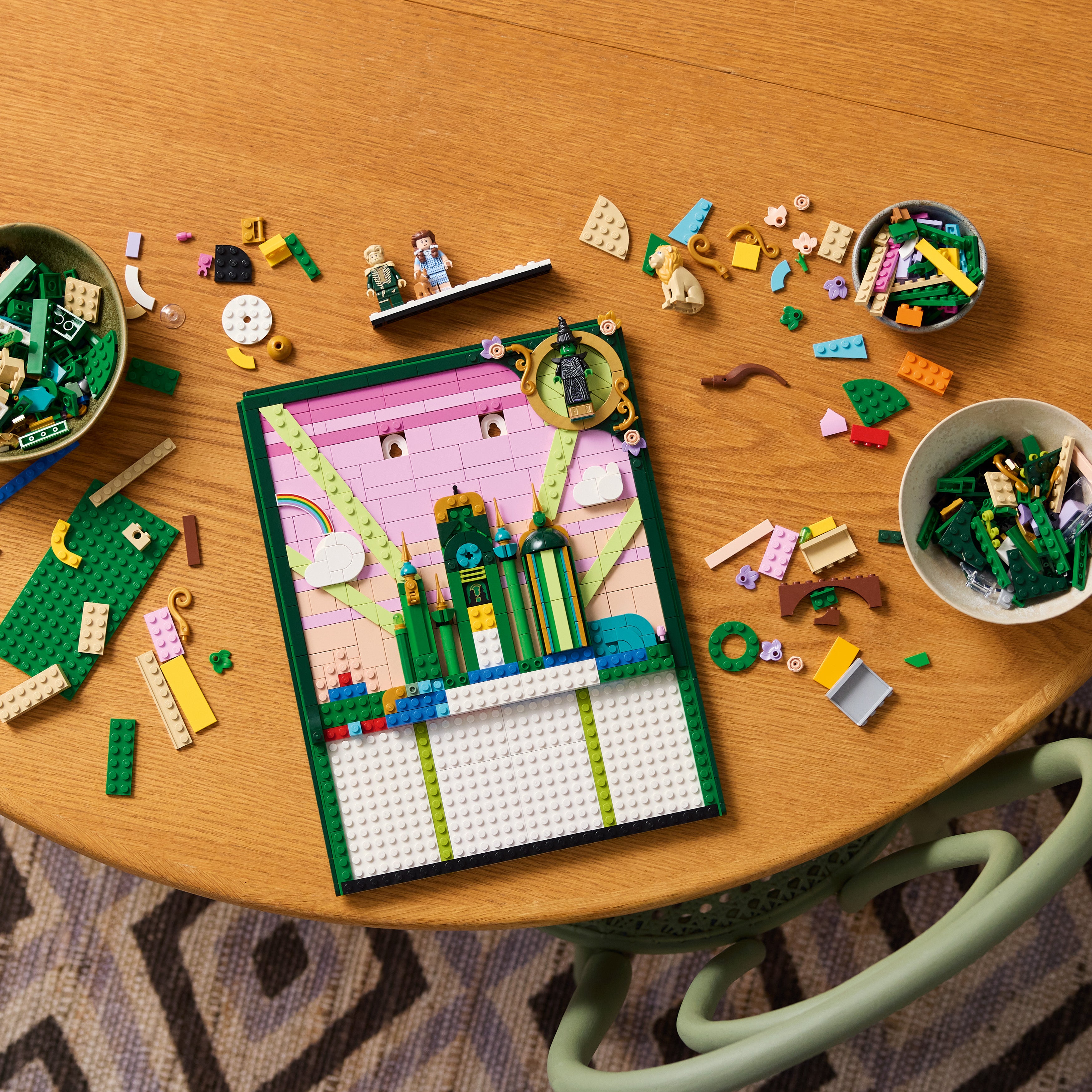 Auf einem Holztisch liegt ein teilweise gebautes LEGO Kunstwerk mit grünen, rosa und weißen Steinen, umgeben von mehreren Schalen mit LEGO Teilen und zwei LEGO Minifiguren.