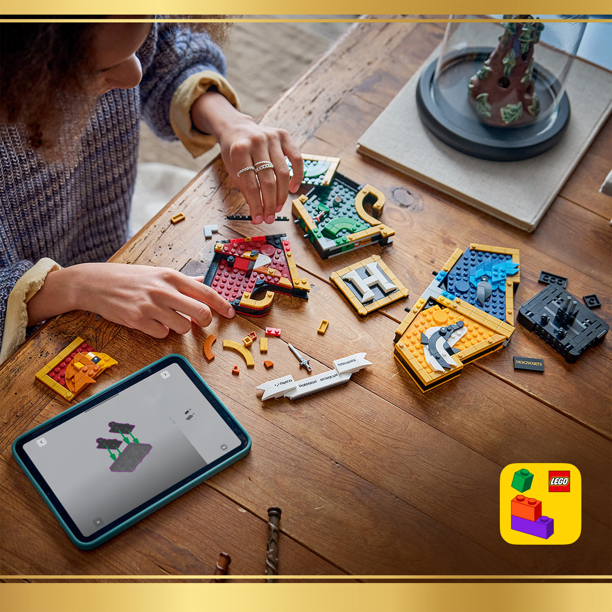 Person baut an einem Tisch mit LEGO Steinen die vier Hogwarts-Hauswappen aus der Harry-Potter-Reihe zusammen, daneben liegt ein Tablet mit Bauanleitung.