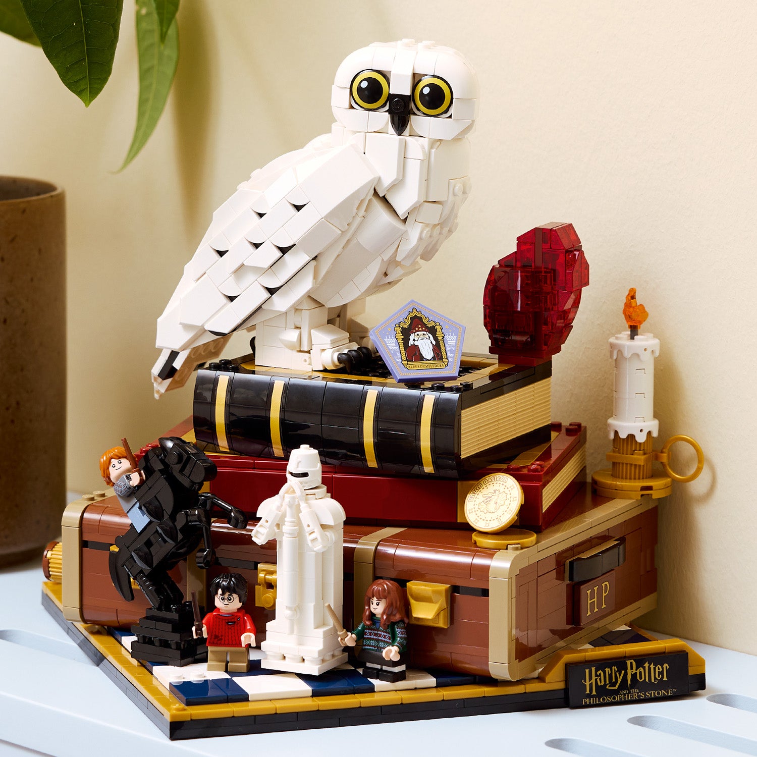 LEGO Harry Potter Sammleredition mit einer weißen Eule auf einem Stapel Bücher, darunter ein roter Edelstein, eine weiße Statue, zwei Minifiguren und ein schwarzes Pferd auf einem Schachbrettmuster, alles auf einem braunen Koffer mit goldenen Details und der Aufschrift 'HP'.