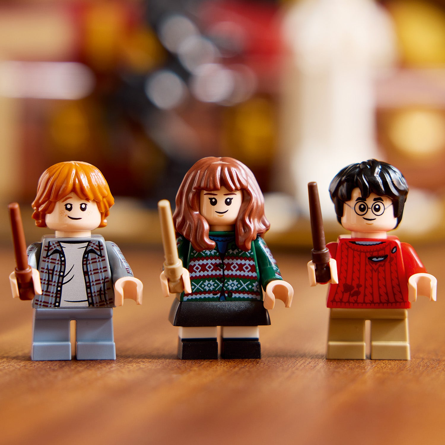 Drei LEGO Harry-Potter-Figuren mit Zauberstäben, darunter eine mit rotem Pullover, eine mit kariertem Hemd und eine mit grün-rotem Norwegerpullover, stehend auf Holzboden vor unscharfem Hintergrund.