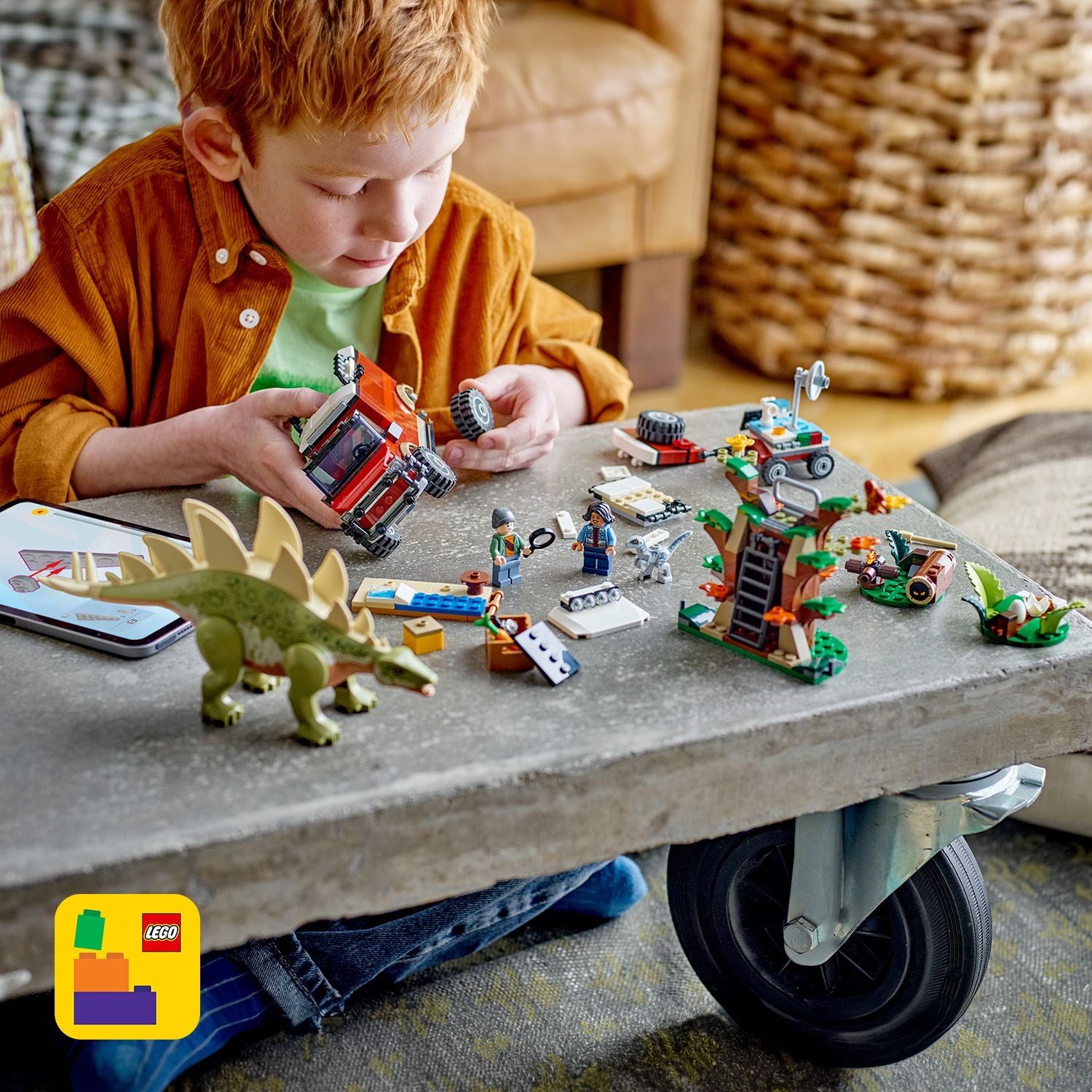 Kind spielt mit LEGO Jurassic World Set auf grauem Tisch, darunter Stegosaurus, Fahrzeug, Baumhaus und mehrere Minifiguren.