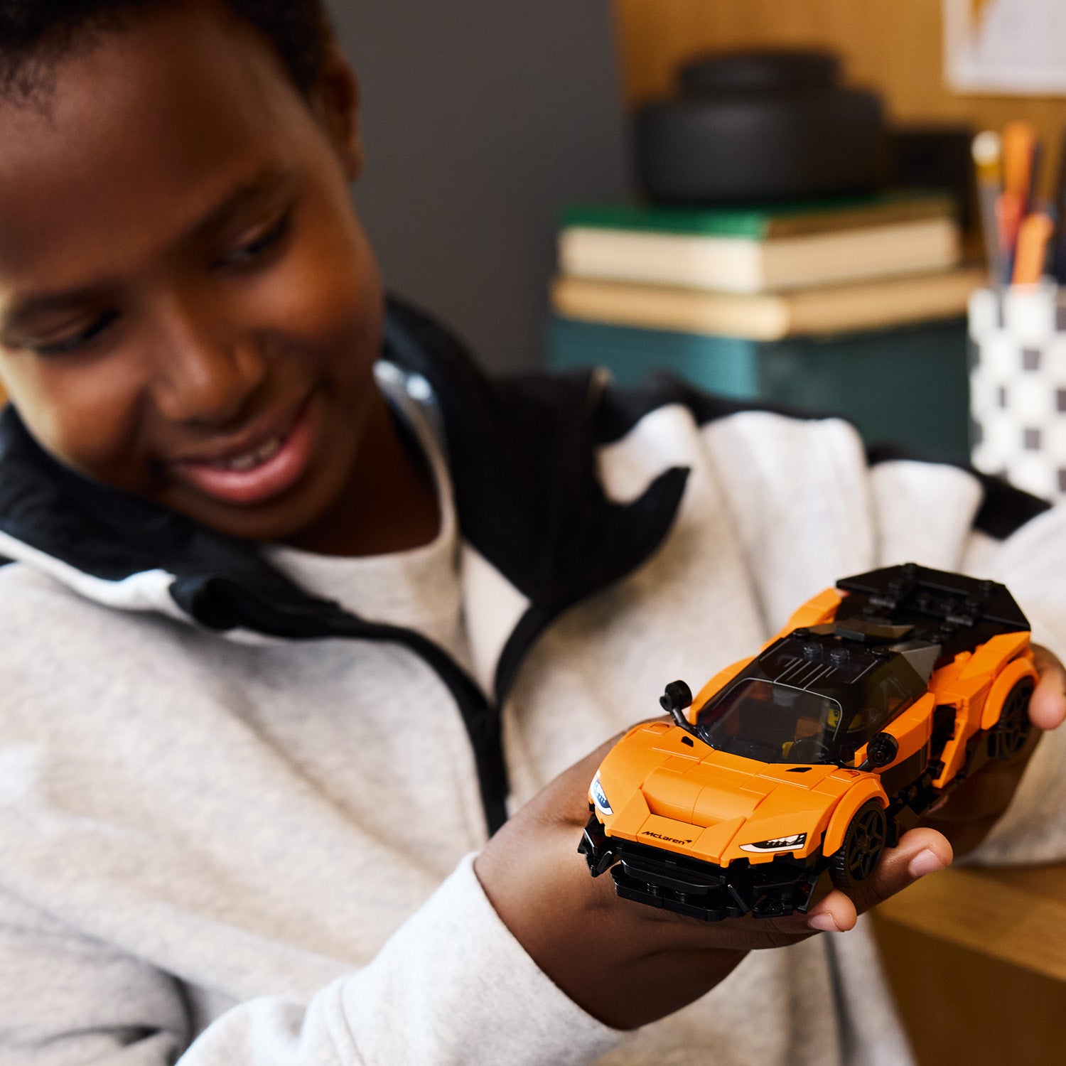 Person hält ein orange-schwarzes LEGO-Modellauto des McLaren Speed Champions in den Händen, im Hintergrund Bücher und Stifte auf einem Tisch.