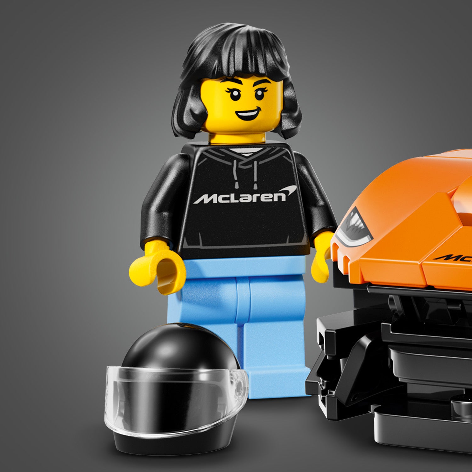 LEGO-Minifigur mit schwarzem McLaren-Hoodie, blauer Hose und schwarzem Helm neben einem orangefarbenen LEGO-McLaren-W1-Modell.