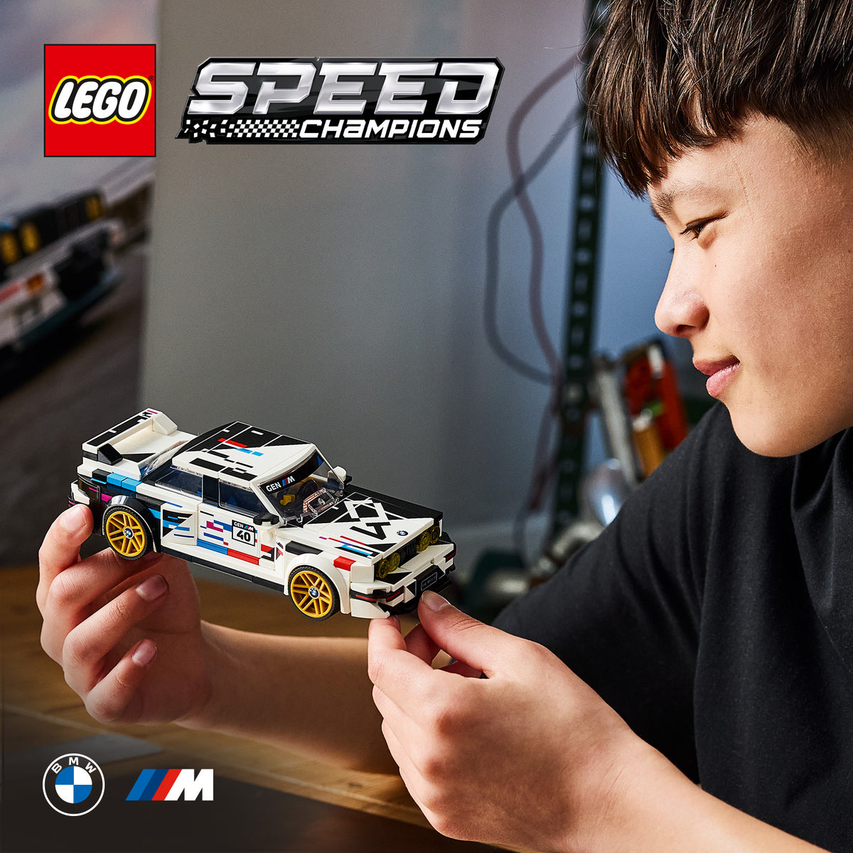 Junge hält ein LEGO Speed Champions Modell des BMW M3 E30 mit weißen, blauen und roten Details und goldfarbenen Felgen in den Händen.