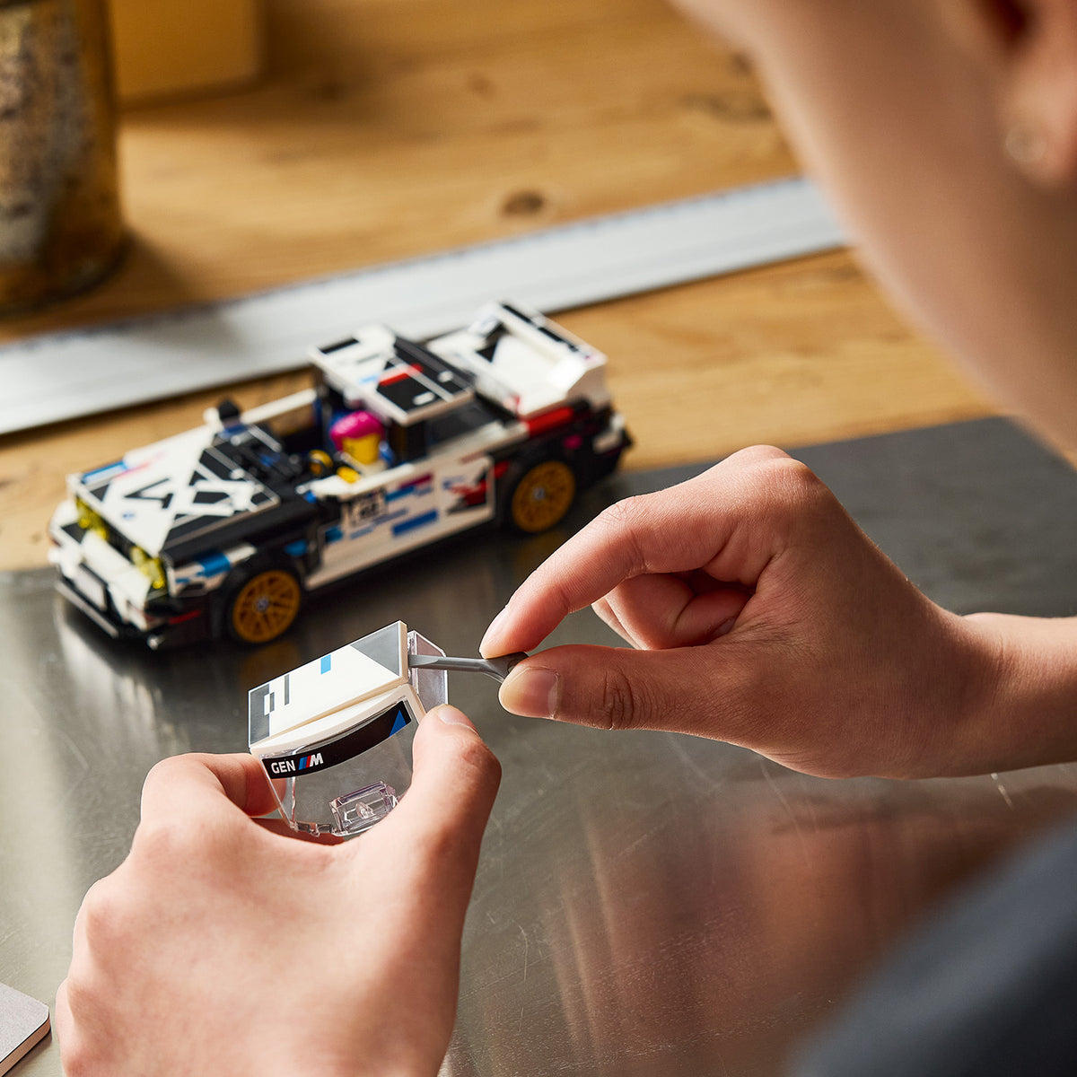 Hände montieren eine transparente LEGO Scheibe mit Aufdruck neben einem zusammengebauten LEGO Modell eines BMW M3 E30 Rennwagens auf einem Tisch