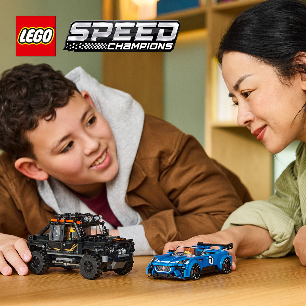 Zwei Personen spielen mit LEGO Speed Champions Modellen eines schwarzen Land Rover Defender und eines blauen Rennwagens auf einem Holztisch